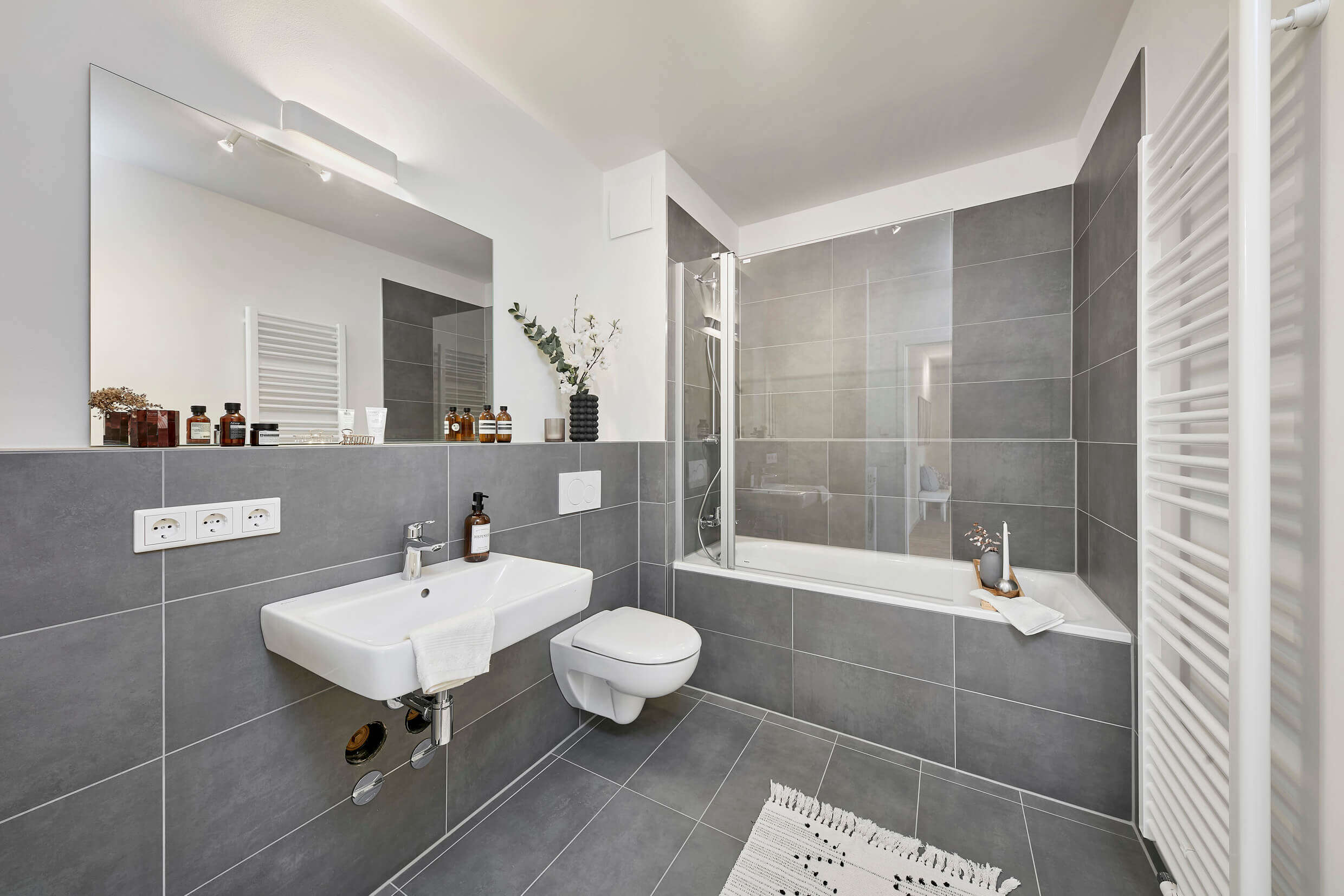 Badezimmer der Musterwohnung zur Miete in der Neuen Meile, graue Wand- und Bodenfliesen, Blick auf das Waschbecken, das WC und die Badewanne.
